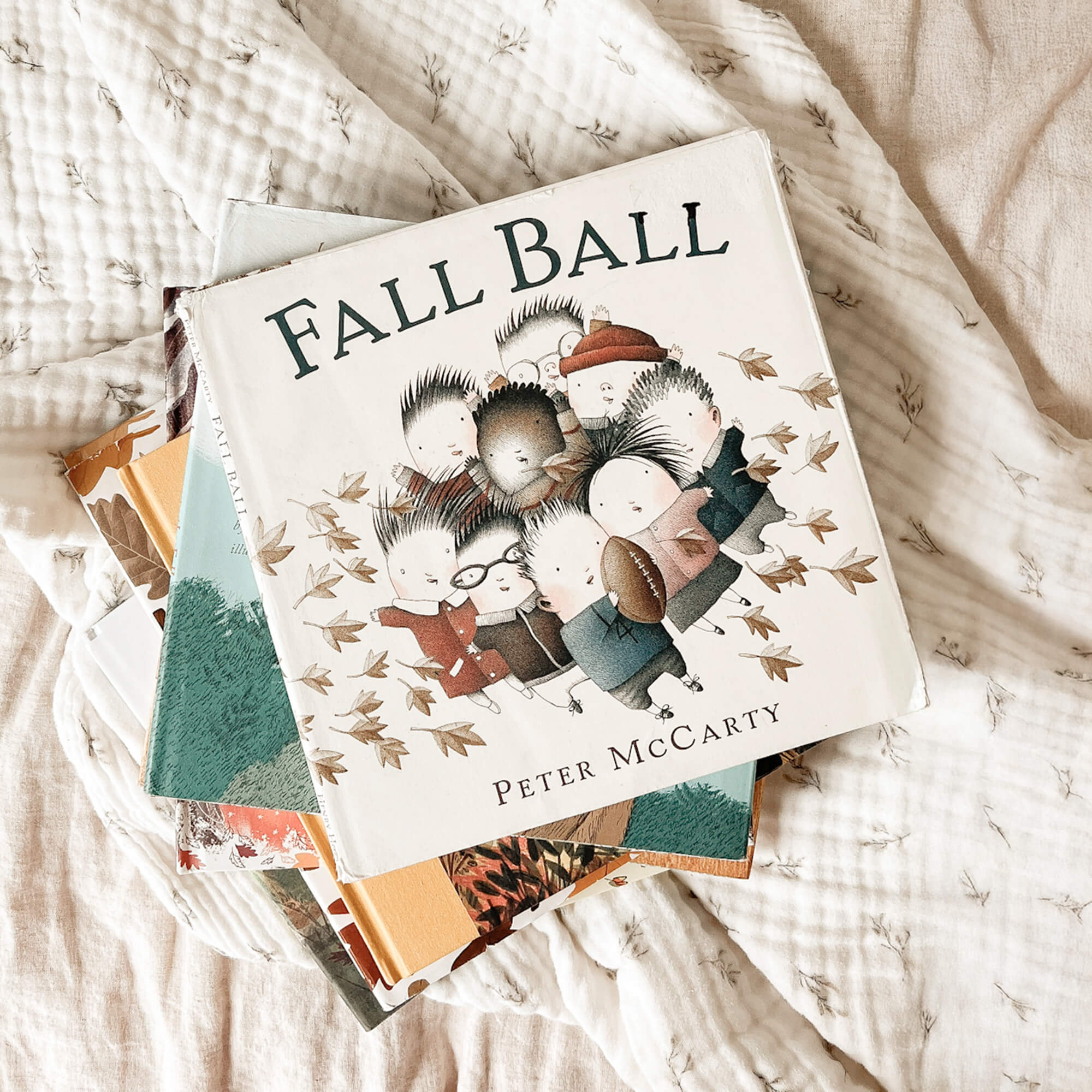 Stack of fall-themed children's books on a muslin blanket with the book "Fall Ball" on top.