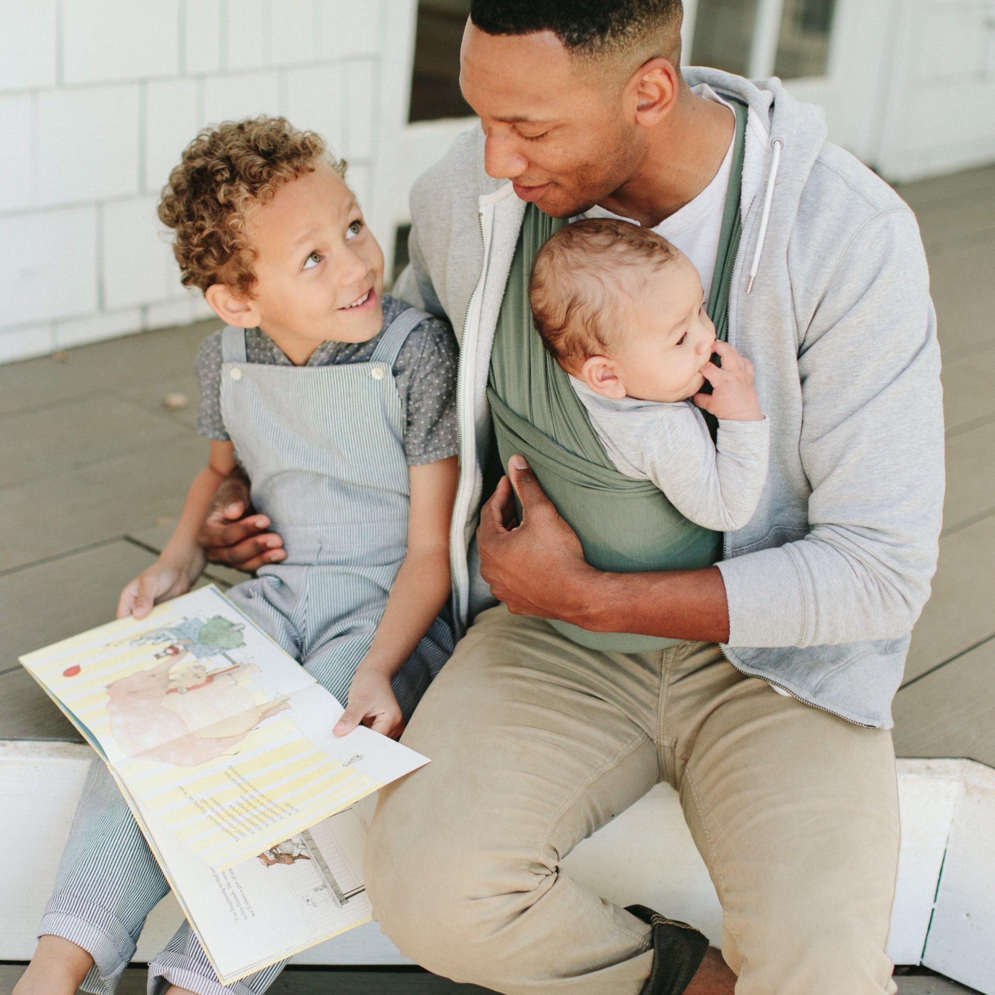 Dad wearing newborn in the Basil Solly Baby Wrap while reading with his older child, showing how babywearing can ease back-to-school chaos by keeping little ones close and hands-free.