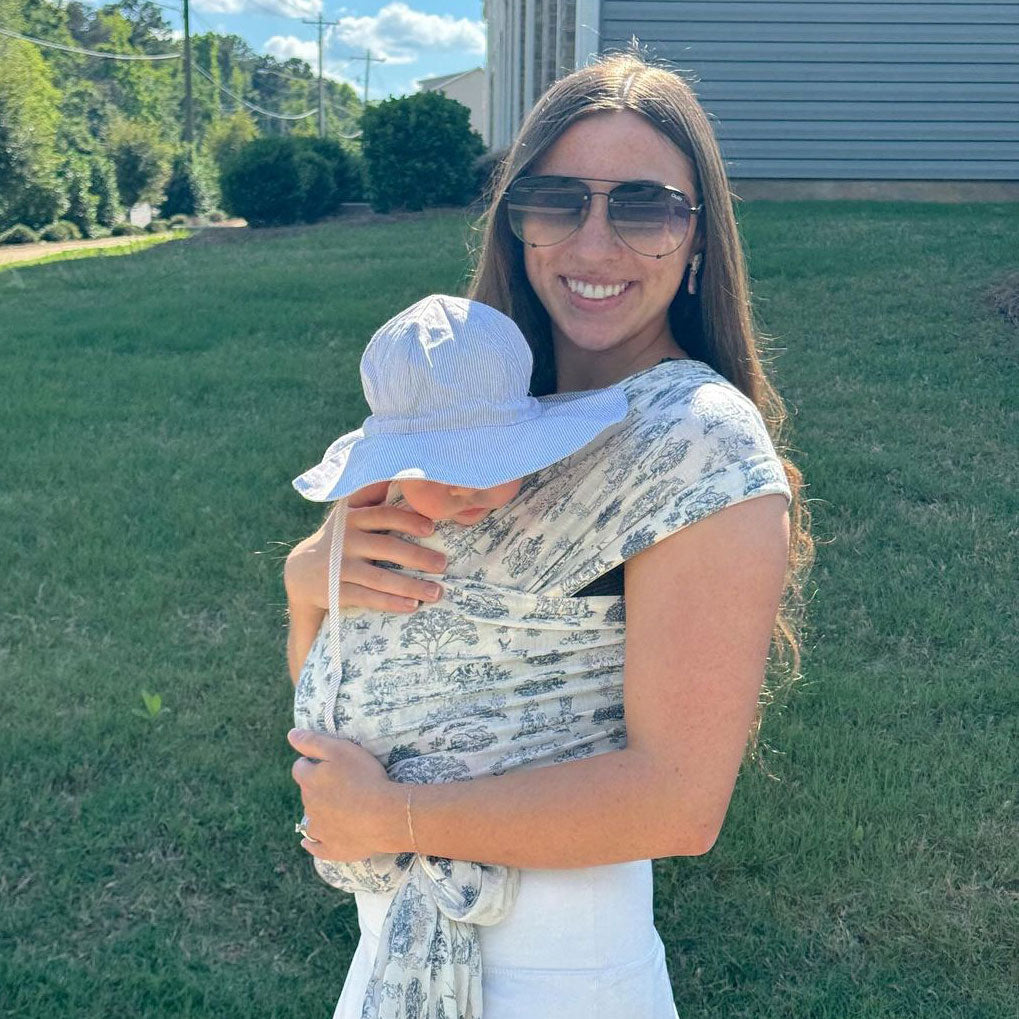 Woman smiles outside on the lawn wearing baby in the Bleu Toile Solly wrap. Baby has wide-brimmed sun hat on.