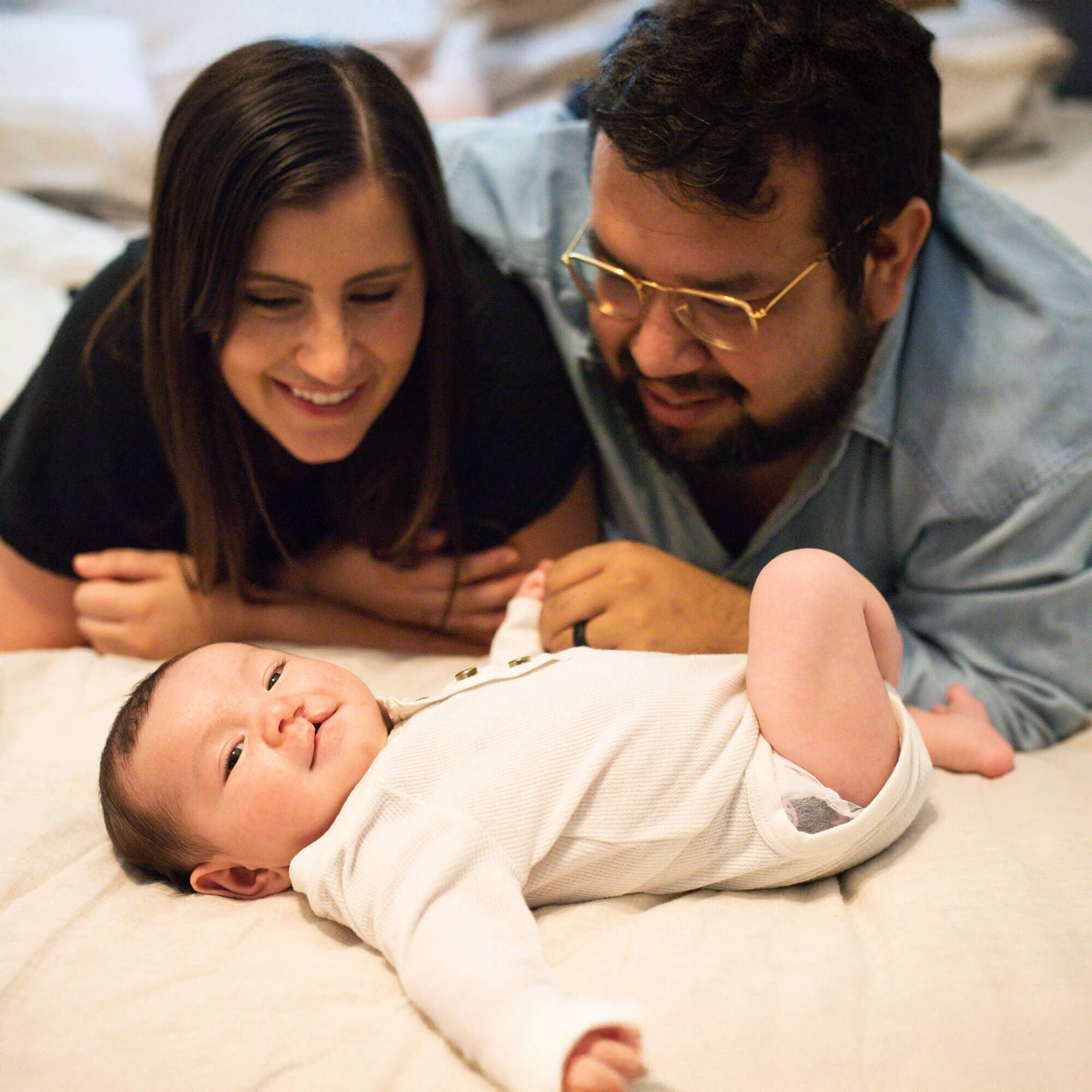 A joyful moment of parents lying beside their baby, who is smiling and dressed in a white onesie. The baby, born with a cleft lip, looks up from a soft bed as both parents gaze at them lovingly.