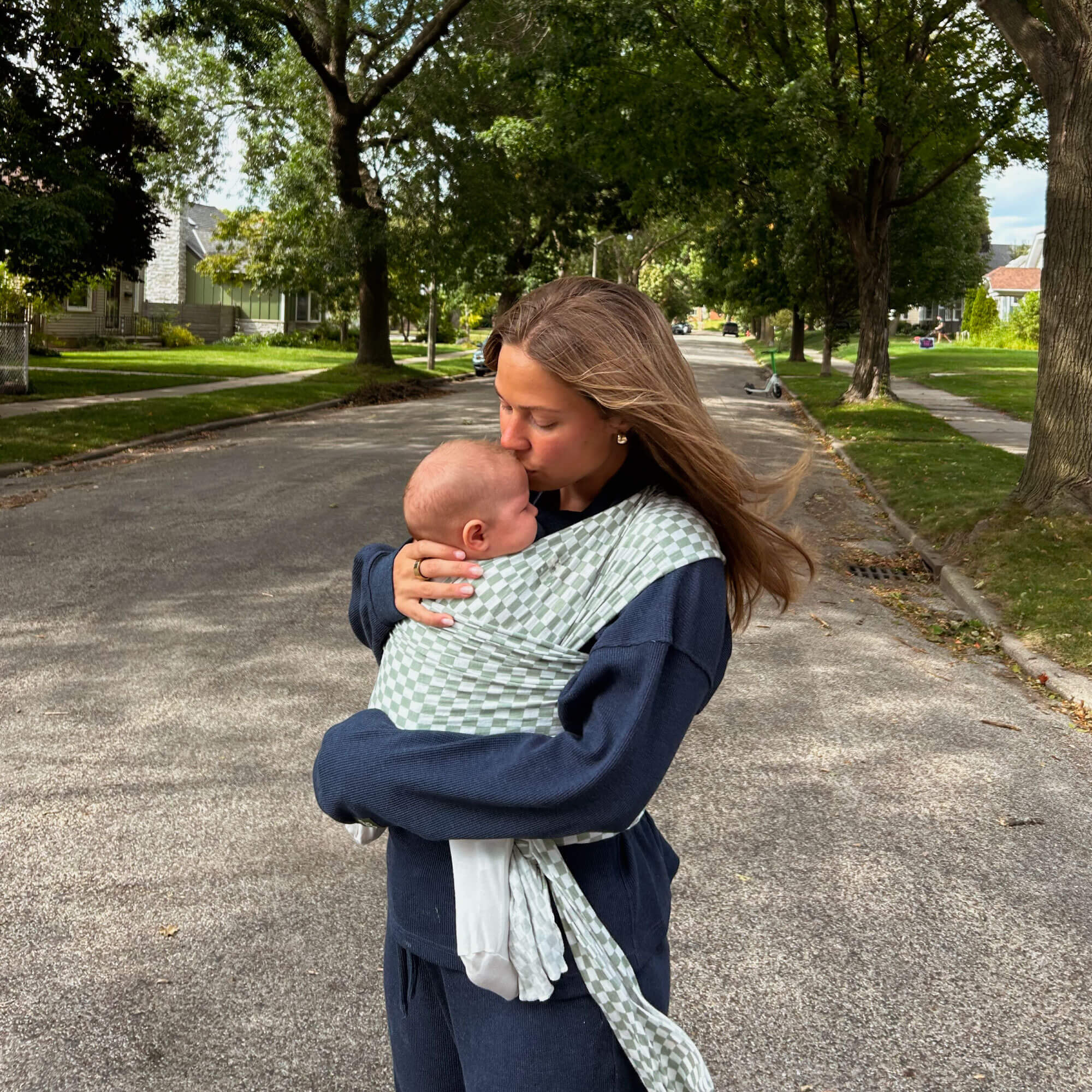 Mom kissing her baby while wearing the Fern Chequer Solly Baby Wrap on a tree-lined street, highlighting love and connection in motherhood.
