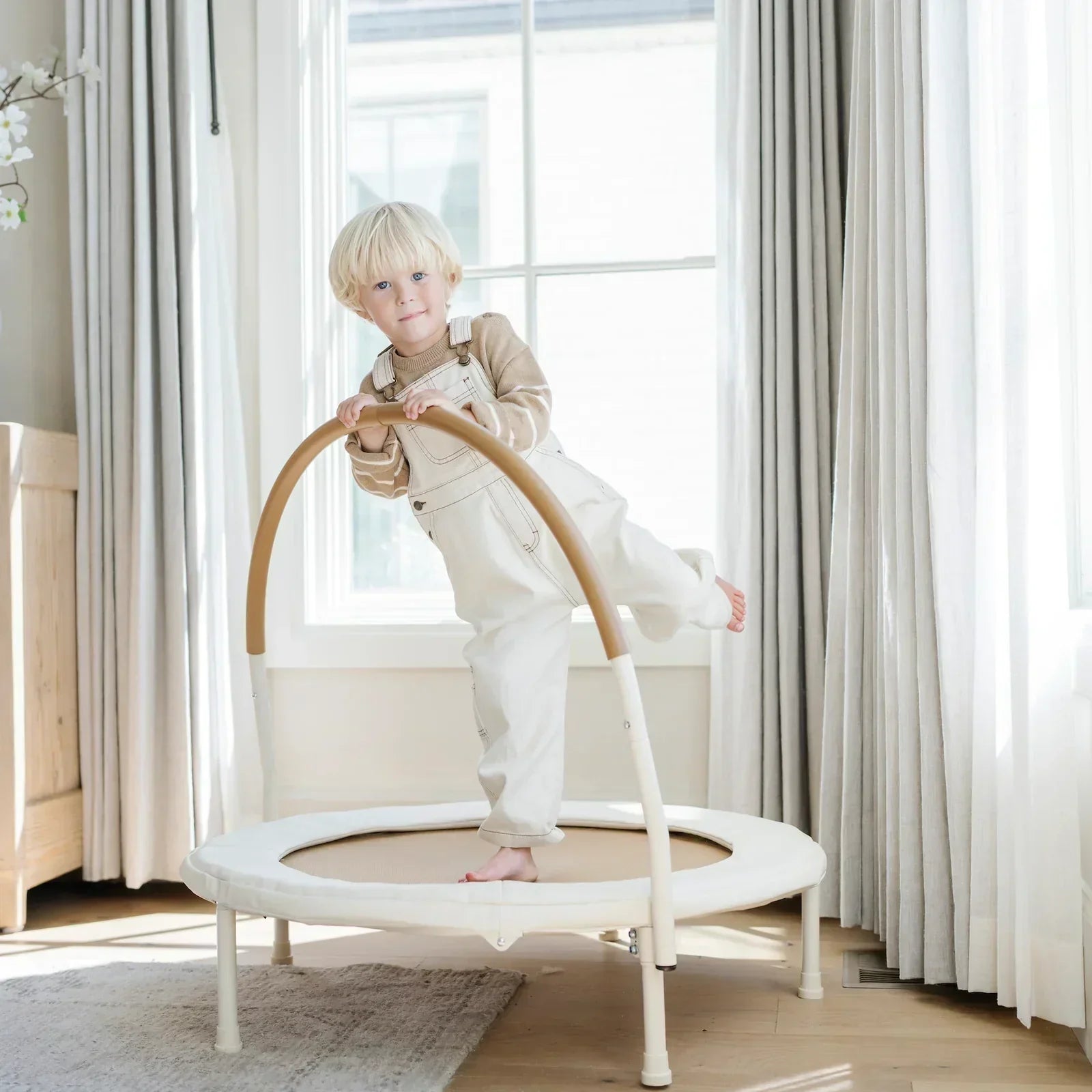 Toddler boy in white overalls standing one one leg on a mini trampoline indoors in a neutral living room