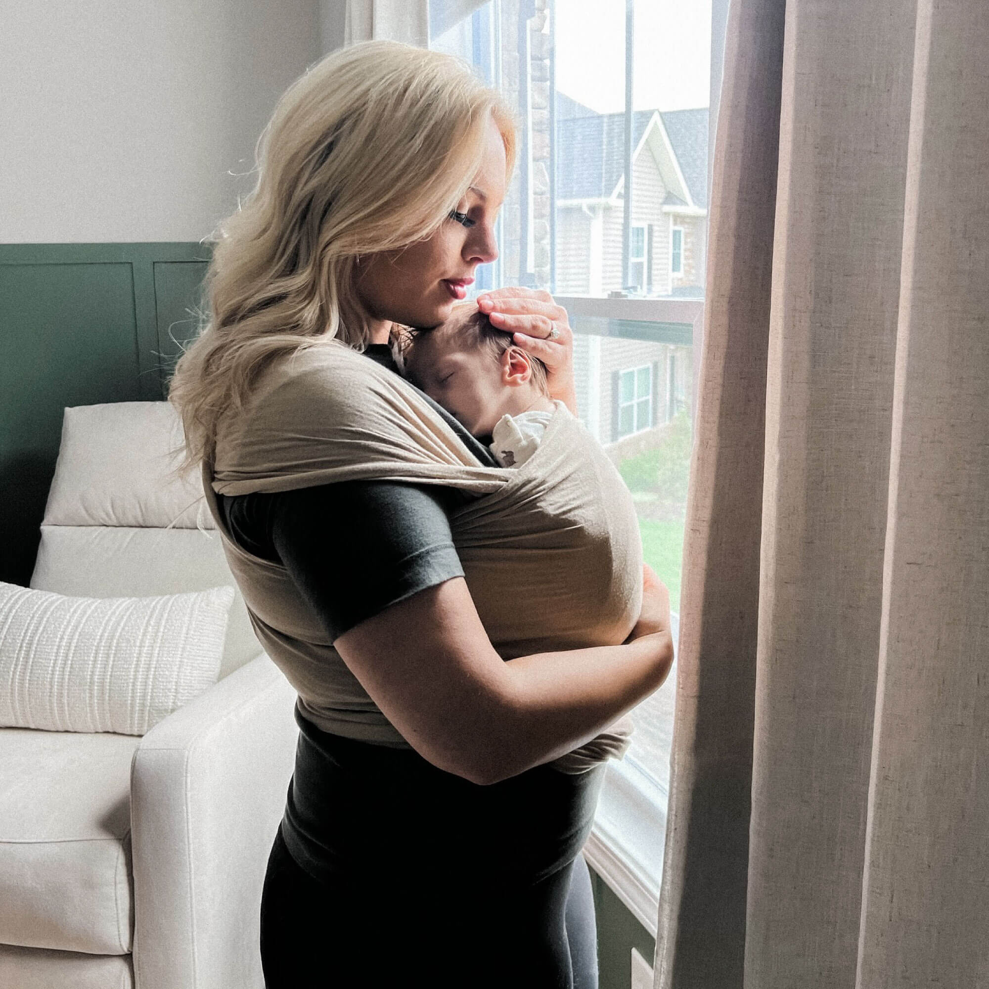 Woman looks out the window while holding baby in the Spelt Baby Wrap carrier