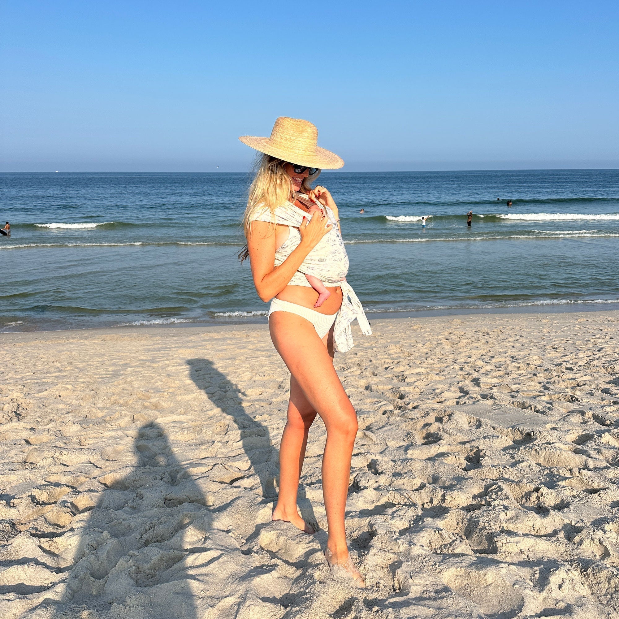 Woman with blonde hair and a straw brimmed hat looks down at baby held close in the Bleu Toile Solly Wrap. Woman is dressed in a white bikini and stands on a sandy beach