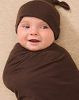 A baby wrapped in a brown swaddle blanket and wearing a matching brown knotted beanie, lying on a textured beige blanket.