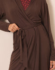 A close-up of a person wearing a dark brown long-sleeve wrap robe with a tie waist, layered over a red patterned top, against a plain light beige wall.