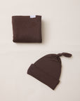 A folded dark brown fabric item with a small white tag is placed above a matching dark brown knotted beanie on a plain warm off-white background.