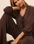 A woman wearing a dark brown long-sleeve jumpsuit with a tie-neck detail, sitting in a relaxed pose against a plain beige background.