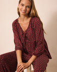 A woman wearing a matching burgundy and red floral print lounge set, sitting on a white stool against a warm beige background.