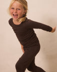 A young blonde girl wearing a dark brown long-sleeve top and matching dark brown leggings, captured in a playful pose against a plain beige wall.