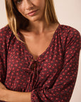 A close-up of a woman wearing a dark pink blouse with a small red and pink floral pattern and a tie detail at the V-neck.