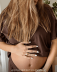 A pregnant woman wearing a brown short-sleeve t-shirt and matching brown bottoms, holding her baby bump with her hands. The photo is taken in a brightly lit indoor setting, and a watermark '@HEALINGWITHHANNAH' is visible at the bottom.