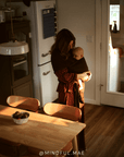 A woman wearing a dark brown and red patterned wrap dress holds a baby in a brown baby wrap carrier inside a warmly lit home kitchen.