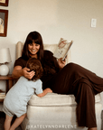 A woman in a dark brown outfit relaxes in a cream armchair, reading a book to a young child who is leaning against the armchair. The scene is set in a cozy, neutral-toned room with a side table and lamp visible.