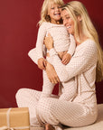 A smiling blonde woman and young child wearing matching cream pajamas with small red stars, sitting together against a dark red wall. A wrapped gift box is visible in the lower left corner.