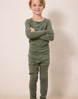 A young boy stands barefoot with arms crossed, wearing an olive green long-sleeve shirt and matching green pants against a plain beige studio background.