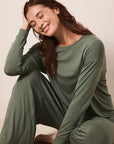 A woman wearing an olive green long-sleeve pajama set with matching pants, sitting on a neutral beige floor against a light beige wall.
