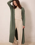 A woman modeling a long sage green open-front cardigan over a beige knit top and matching wide-leg pants, standing against a plain beige studio backdrop.