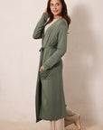 A woman modeling a long-sleeve sage green robe with a tie waist and front pockets, worn over a cream top and beige pants, standing against a plain beige studio wall.