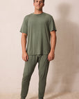 A man modeling a matching two-piece pajama set in olive green, consisting of a short-sleeve crewneck t-shirt and matching jogger pants, standing against a plain beige wall.