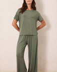 A woman modeling a matching two-piece lounge set in sage green, featuring a short-sleeve crewneck top and wide-leg pants, standing against a plain beige wall.