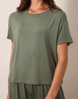 A woman wearing a loose, short-sleeve crewneck t-shirt in sage green, paired with matching sage green pants, photographed against a neutral beige background.