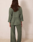 A woman viewed from behind wearing an olive green two-piece lounge set consisting of a long-sleeve peplum top and matching wide-leg pants, standing barefoot against a plain beige wall.
