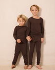 Two young boys standing barefoot against a plain beige studio wall, wearing matching dark brown long-sleeve crewneck pajamas or shirts and pants.