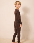A young boy with blonde hair stands in profile wearing a matching dark brown long-sleeve top and jogger pants set, barefoot against a plain beige studio backdrop.