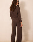 A woman wearing a matching dark brown knit lounge set, consisting of a long-sleeve top and wide-leg pants, shown from a back-three-quarter view against a plain beige studio background.