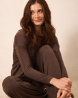 A woman with long brown hair sits cross-legged on a light beige textured floor against a neutral beige wall, wearing a matching long-sleeve crewneck top and wide-leg pants in a solid dark brown color.