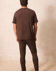 A man viewed from behind wearing a matching dark brown short-sleeve t-shirt and long pants pajama set, standing barefoot against a plain beige studio background.