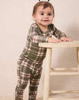A toddler standing barefoot and leaning on a light wooden stool, wearing a long-sleeve plaid pajama set in muted green, beige, and burgundy tones against a plain cream backdrop.