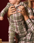 A baby wearing a matching plaid jumpsuit sits on a woman's lap. The baby's outfit is olive green with a red and beige plaid pattern.