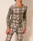 A child wearing a matching long-sleeve top and pants pajama set in an olive green plaid pattern with beige and white stripes, set against a plain beige wall.