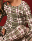 A close-up of a child wearing a long-sleeve plaid pajama top and matching pants in muted green, beige, and burgundy tones, seated against a dark red background.