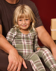 A young blonde girl wearing a green and red plaid pajama set sits on her father's lap against a dark red background.