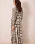 A woman wearing a matching long-sleeve plaid pajama set in sage green, cream, and white, shown from a side-back angle against a plain beige studio backdrop.