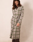 A woman modeling a long-sleeve plaid bathrobe in sage green, cream, and brown, standing against a plain beige studio background.