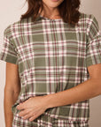 A woman wearing a short-sleeve plaid top in sage green, cream, and burgundy, shown from the shoulders to the waist against a plain beige background.