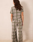 A woman shown from the back wearing a short-sleeve plaid jumpsuit in olive green, beige, and white, standing against a plain beige wall.