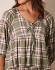 A woman wearing a green plaid short-sleeve top with a tie neckline, shown in a close-up shot against a neutral beige background.