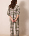 A woman stands barefoot against a plain beige wall, viewed from the back, wearing a green plaid pajama set with a long-sleeve top and matching pants.
