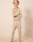 A young boy with blonde hair stands barefoot against a warm beige studio backdrop, wearing a matching beige long-sleeve top and jogger-style pants set.