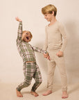 Two children modeling long-sleeve pajamas in a studio setting. The child on the left wears a green, pink, and white plaid pajama set and is dancing joyfully while holding hands with the child on the right, who wears a solid beige pajama set.
