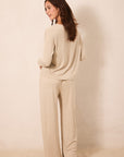 A woman stands barefoot against a plain beige wall, wearing a matching beige knit lounge set consisting of a long-sleeve top and wide-leg pants.