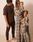 A family of five wearing matching plaid pajamas stands together against a plain beige studio background. The father wears a solid dark brown pajama set, while the mother, baby, and two children wear green, beige, and brown plaid long-sleeve pajama sets.