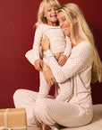 A smiling blonde woman and young blonde child wearing matching cream pajamas with small red polka dots, sitting together against a dark red background with a wrapped gift box on the floor.