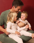 A father and two children wearing matching light red and white twinkle star pajamas, sitting together against a solid dark red background.