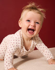 A baby crawling on a beige fabric surface against a solid dark red background, wearing a cream-colored long-sleeve bodysuit with a small red star pattern.